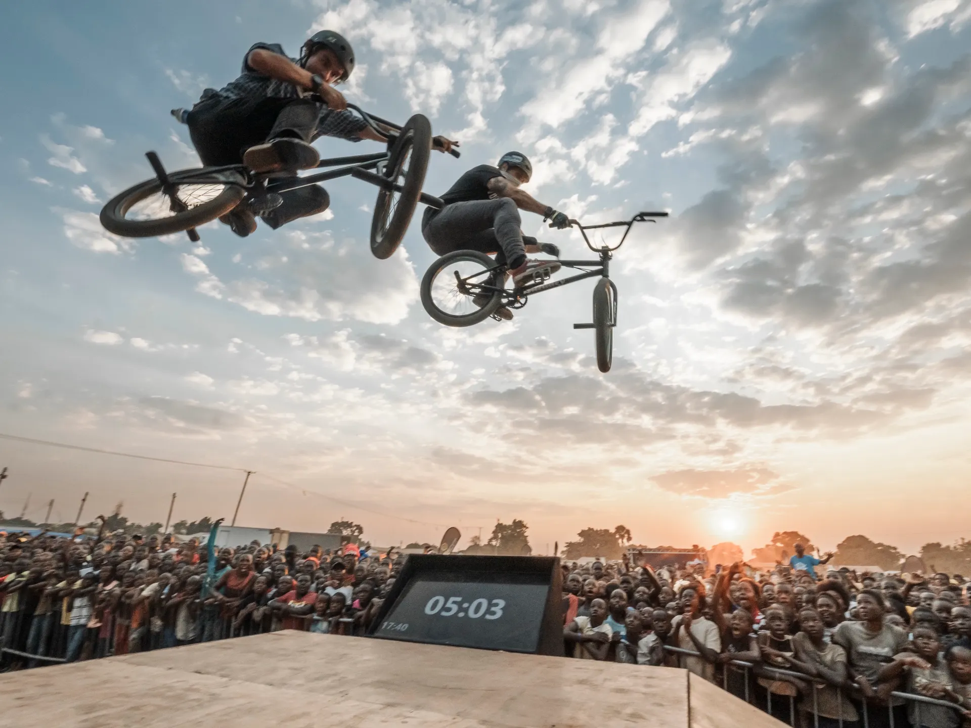 Two StuntDudes riders mid-backflip over the ramp at sunset above a packed crowd